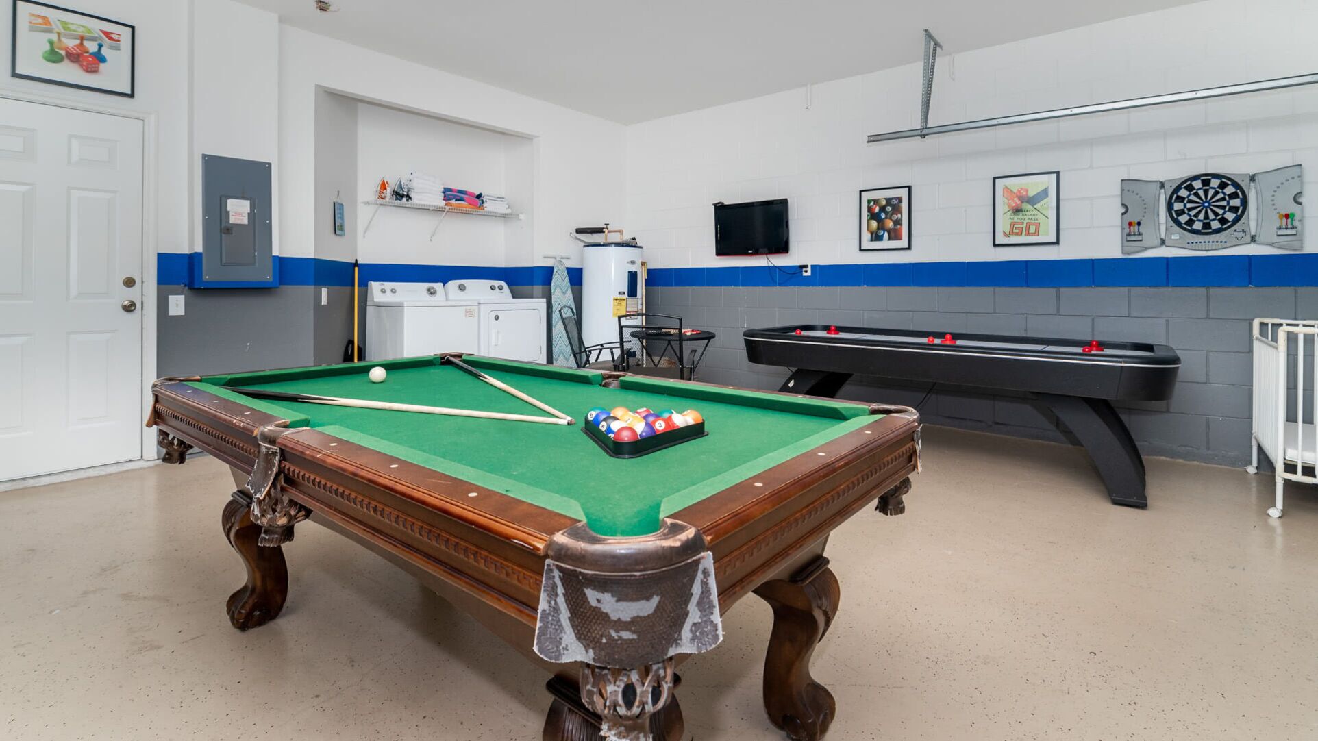 Une table de billard avec des billes, une queue de billard et un porte-billes. Il y a un téléviseur fixé au mur et un jeu de fléchettes.