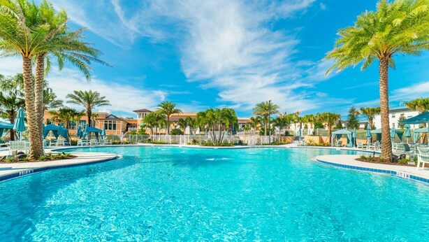 A large outdoor swimming pool surrounded by palm trees and lounge chairs, with buildings in the background.