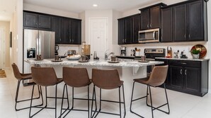 A modern kitchen with dark cabinetry, a central island with bar stools, stainless steel appliances, and a white tiled backsplash.