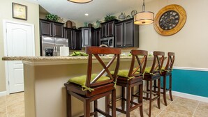 Une cuisine avec des meubles de couleur bois foncé, un réfrigérateur en acier inoxydable et un coin bar doté de tabourets en bois et d’un coussin vert.