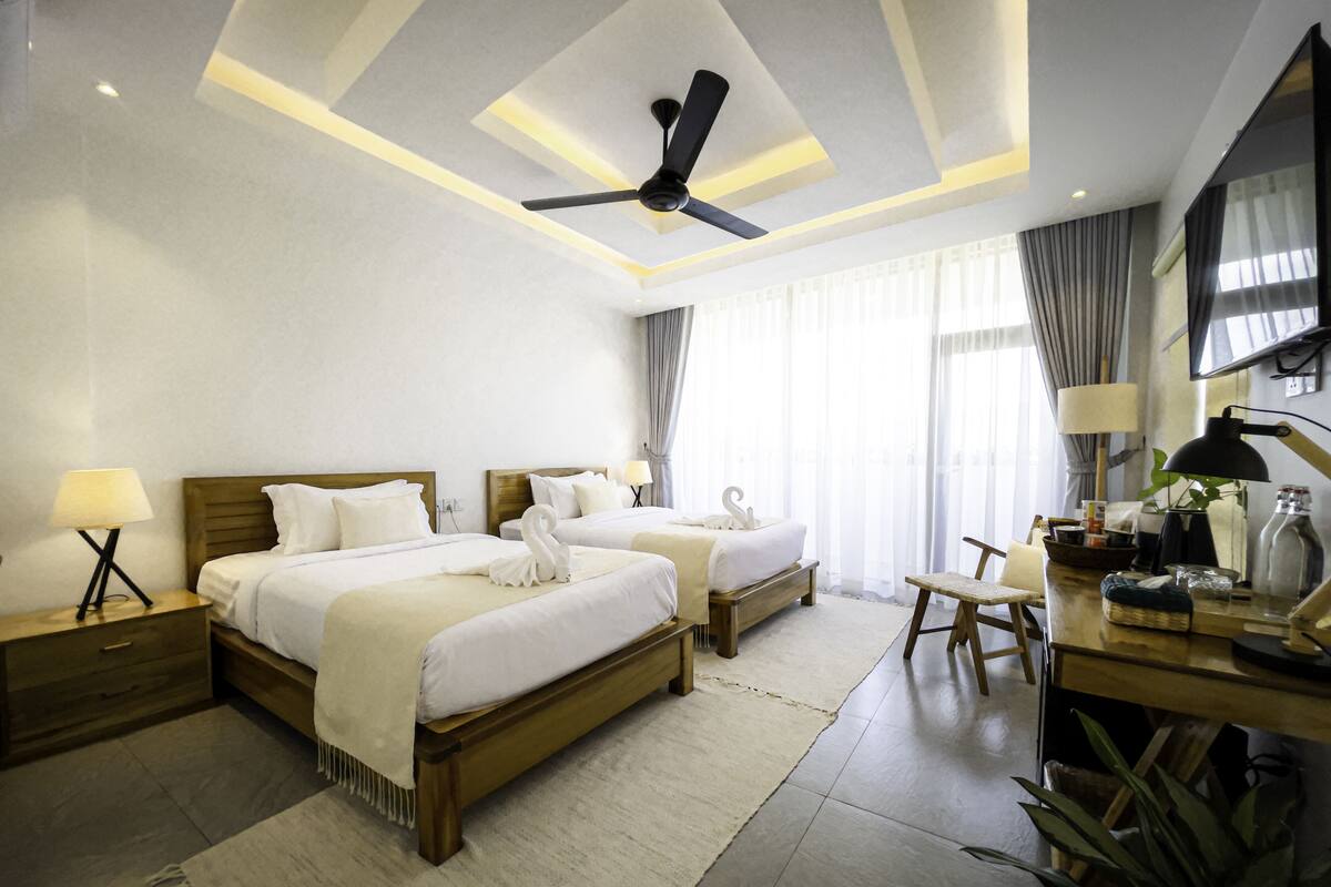 A hotel room with two beds, a desk, a chair, a TV, and a ceiling fan.