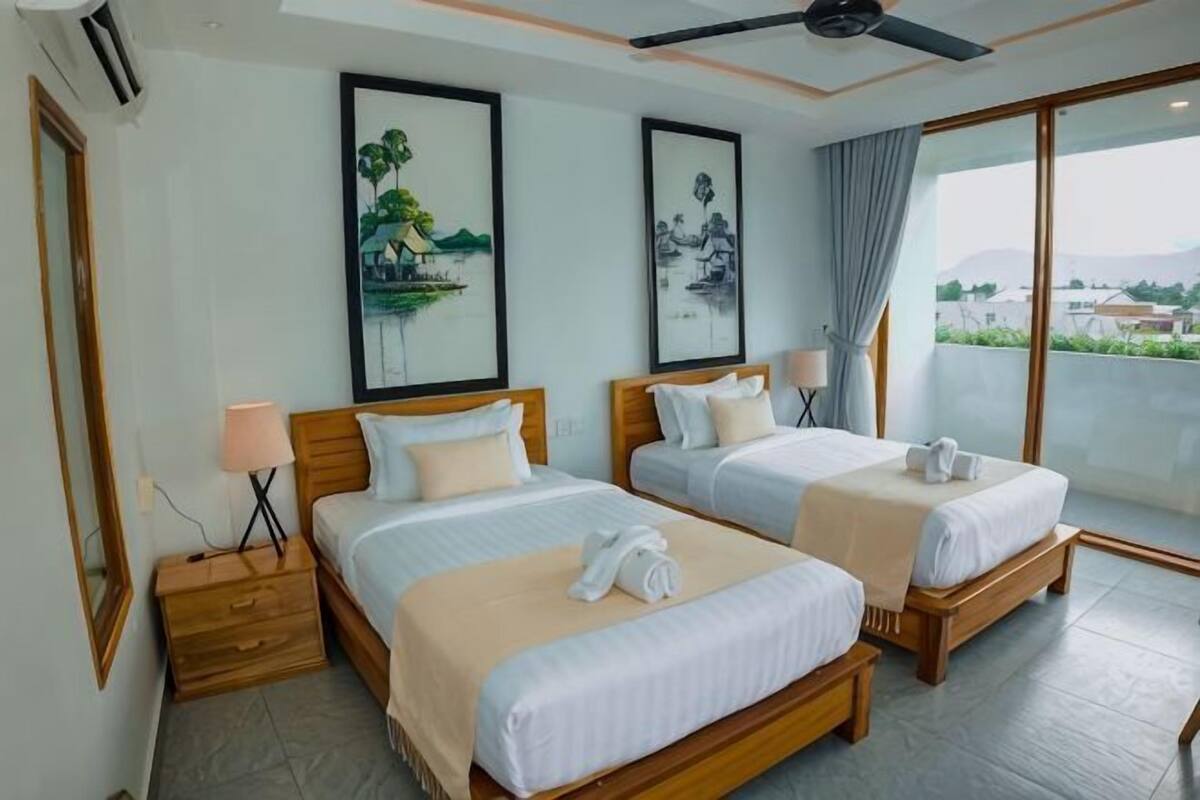 A hotel room with two beds, a ceiling fan, artwork on the walls, and a view through large windows.