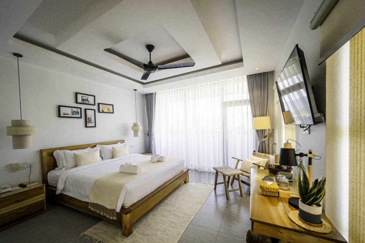 A modern hotel room with a large bed, a desk with a lamp, a television, and a ceiling fan.