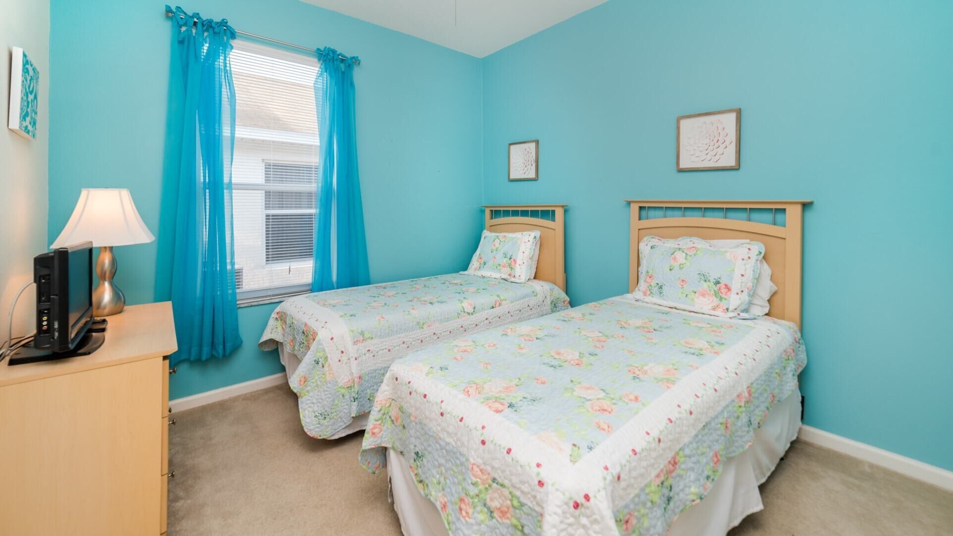 A room with two beds, a desk with a lamp, and a window with blue curtains.