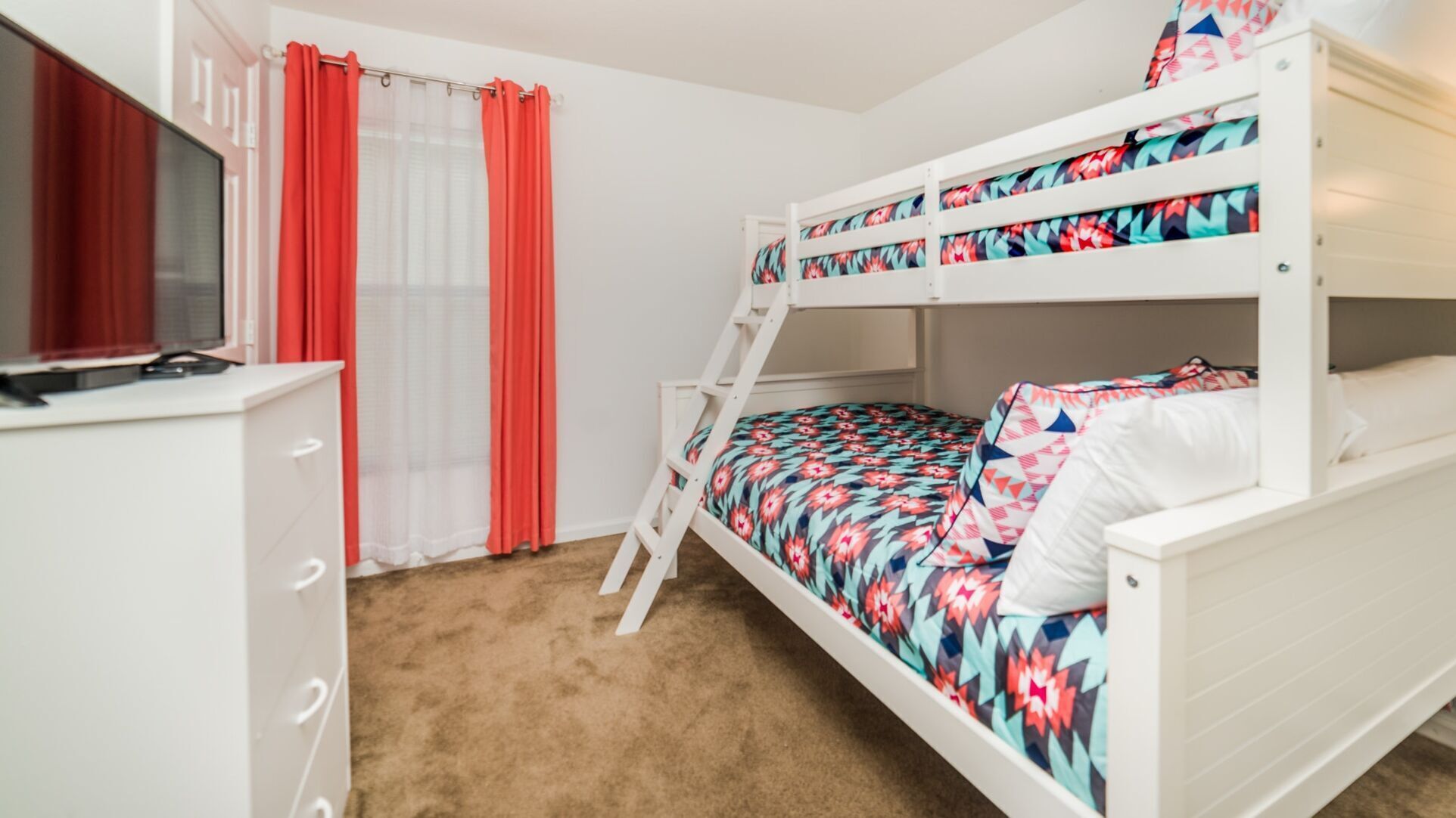 A bunk bed with a colorful bedspread, a white dresser with drawers, and a window with red curtains.
