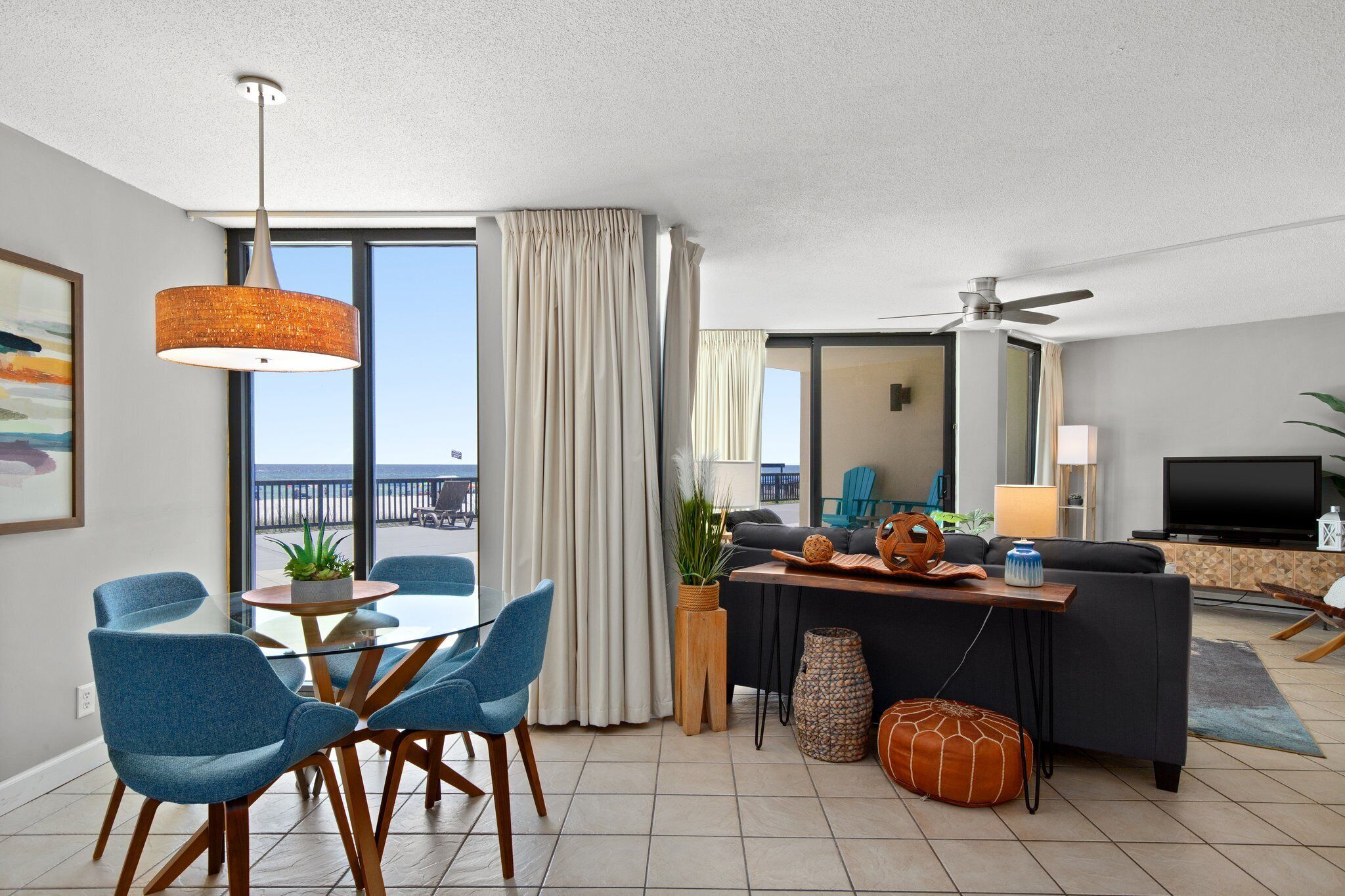 A modern living room with a dining area, large windows, a ceiling fan, and a view of the ocean.