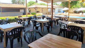 An outdoor seating area with tables and chairs, a pool in the background, and a covered roof structure. - Pousada Vila Cajuína - Luís Correia (Luis Correia)