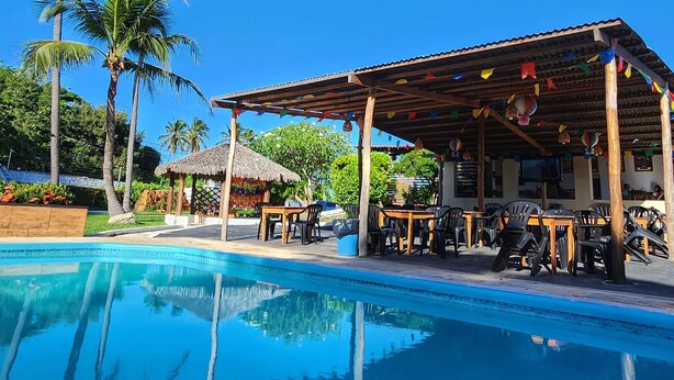 A poolside area with a covered seating area, palm trees, and a thatched-roof structure. - Pousada Vila Cajuína - Luís Correia (Luis Correia)