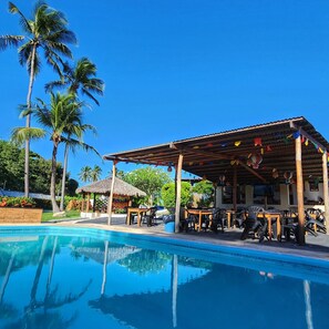 A poolside area with a covered seating area, palm trees, and a thatched-roof structure. - Pousada Vila Cajuína - Luís Correia (Luis Correia)