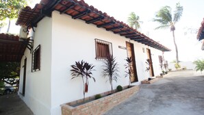 A single-story building with a tiled roof, a brick foundation, and a white exterior wall. There are potted plants along the front and a sign on the wall. - Pousada Vila Cajuína - Luís Correia (Luis Correia)