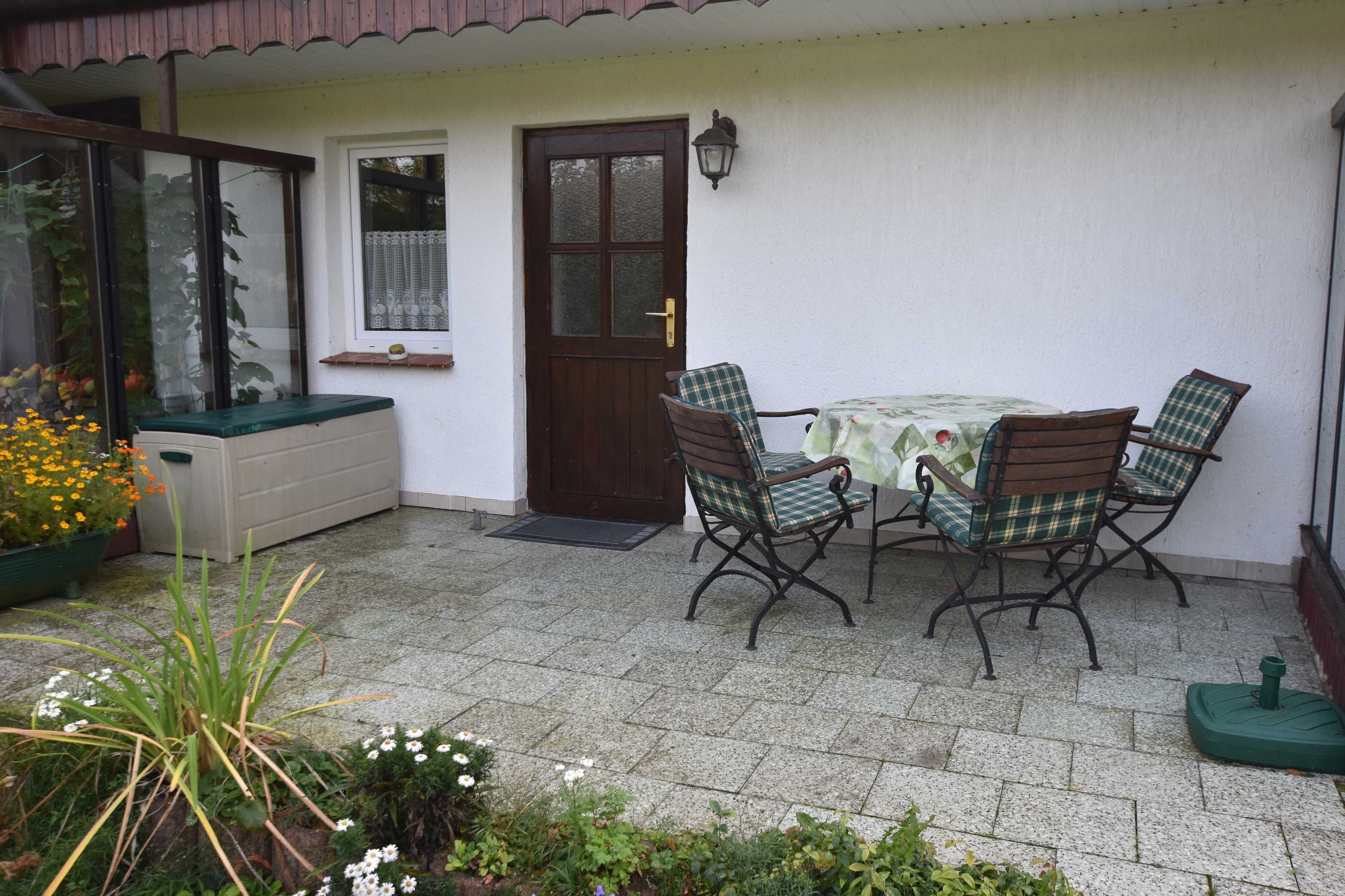 Eine Terrasse mit Tisch und vier Stühlen, einer Tür und einem Fenster mit Vorhang.