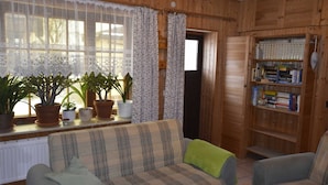 A cozy room with wooden walls, a plaid sofa, a bookshelf, and several potted plants near a window with lace curtains.