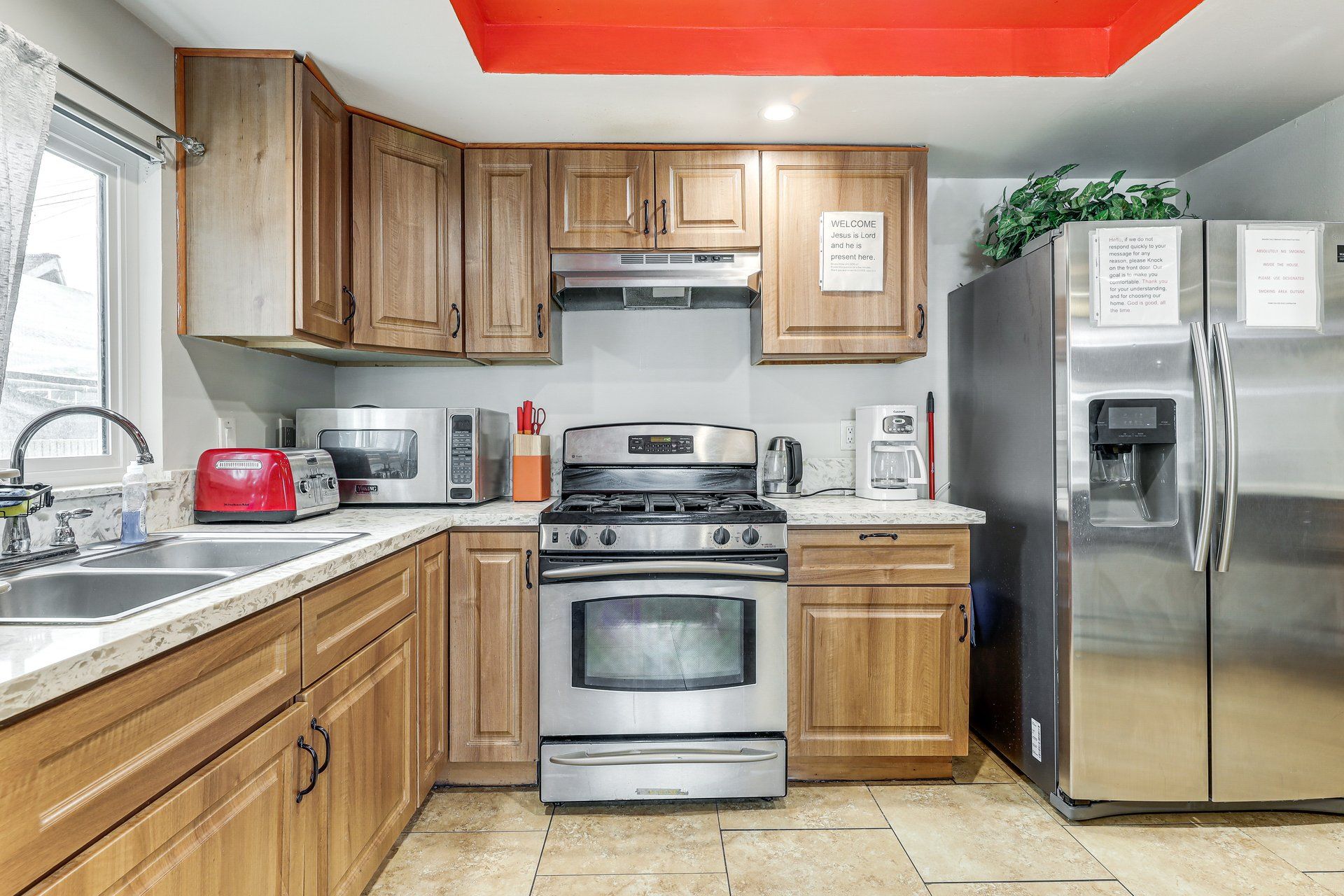 A modern kitchen with wooden cabinets, stainless steel appliances including a refrigerator, oven, and microwave, and a red ceiling.