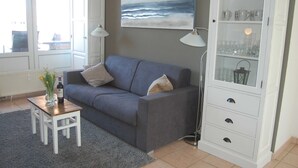 A living room with a grey sofa, a small wooden table with a bottle and flowers, a floor lamp, and a cabinet with glassware. - Ferienwohnung Rerik - Ferienwohnung (Rerik)