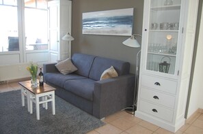 Ein Wohnzimmer mit grauem Sofa, einem kleinen Holztisch mit Flasche und Blumen, einer Stehlampe und einem Schrank mit Gläsern.