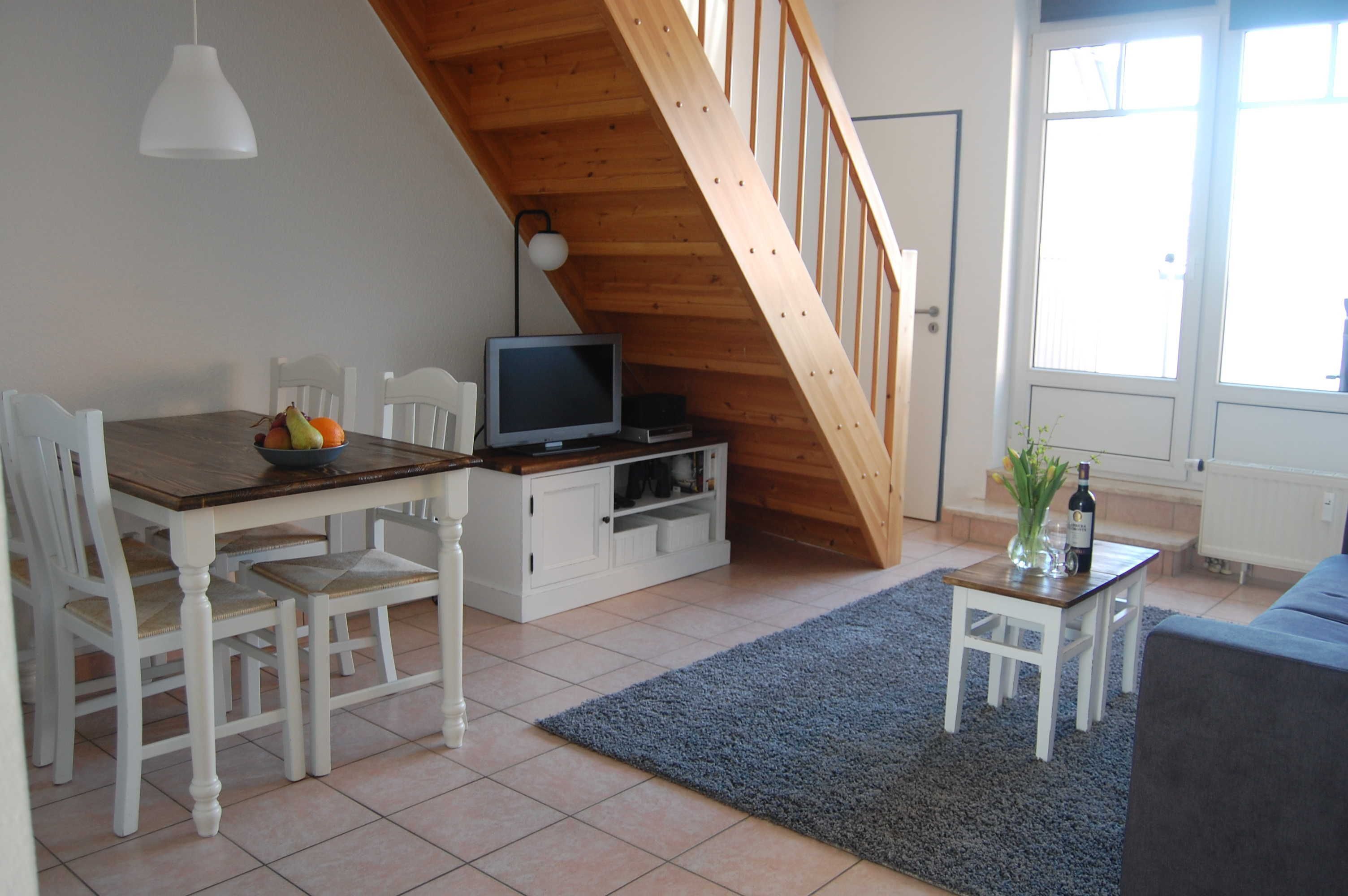 Un comedor con una mesa y sillas de madera, un televisor sobre un mueble, y una escalera que sube a un nivel superior.