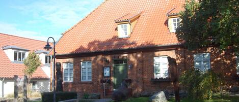 En traditionell byggnad i tegel med ett tak täckt av takpannor, en grön dörr och en liten trädgårdsyta med en skulptur i sten.