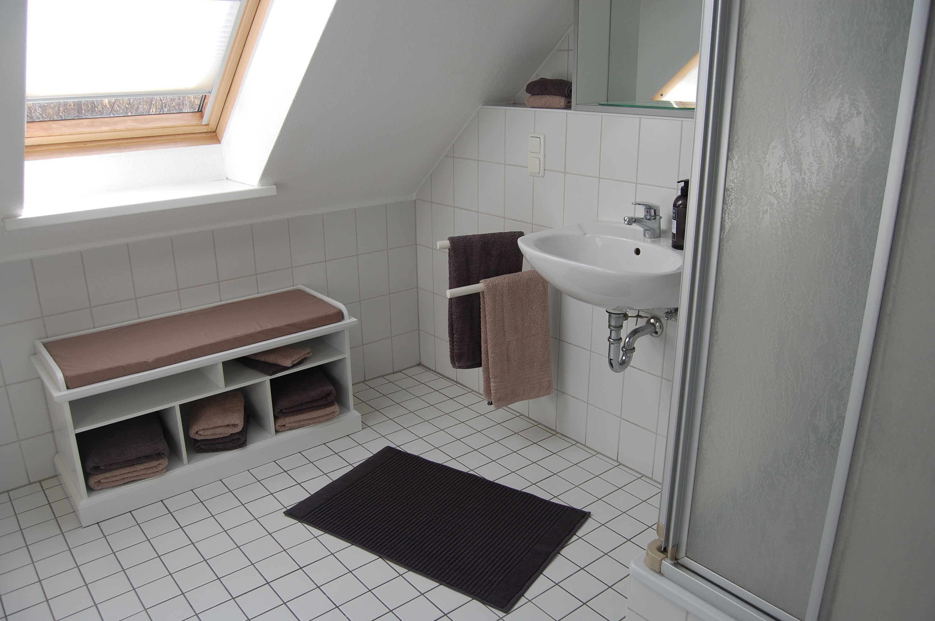 Ein kleines, modernes Badezimmer mit Oberlicht, einem weißen Waschbecken, einer Sitzbank mit Handtüchern und einem dunklen Teppich auf dem Boden.