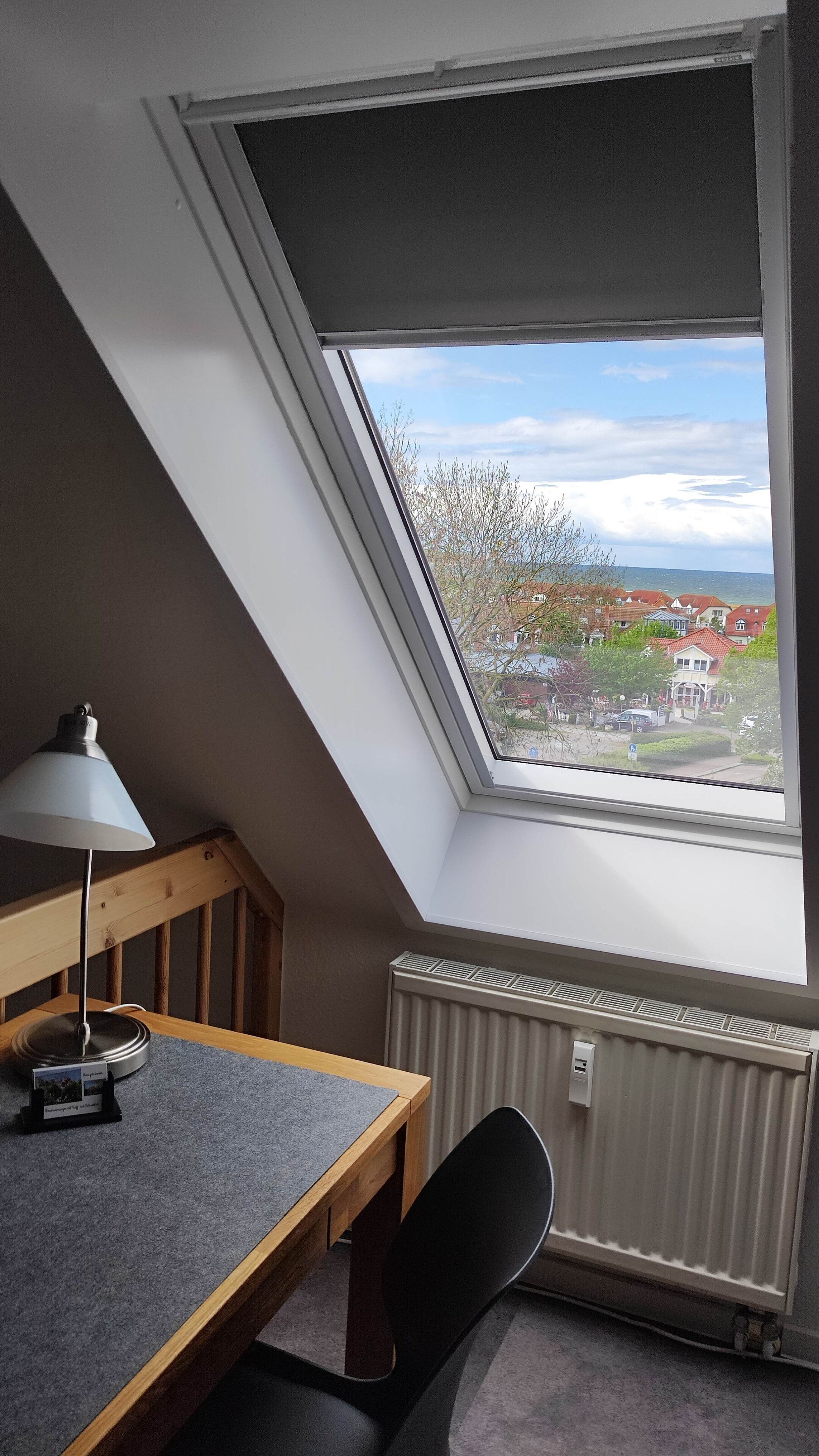 A room with a sloped ceiling, a table with a lamp, a chair, and a view of a residential area through a window.