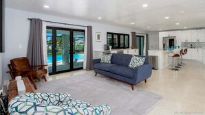 Un salon moderne avec un canapé, un pouf à motifs, une chaise en bois et une vue sur une piscine, visible à travers de grandes portes vitrées.