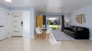 Un salon moderne avec un canapé noir, une table à manger avec des chaises et une vue à travers une porte vitrée.