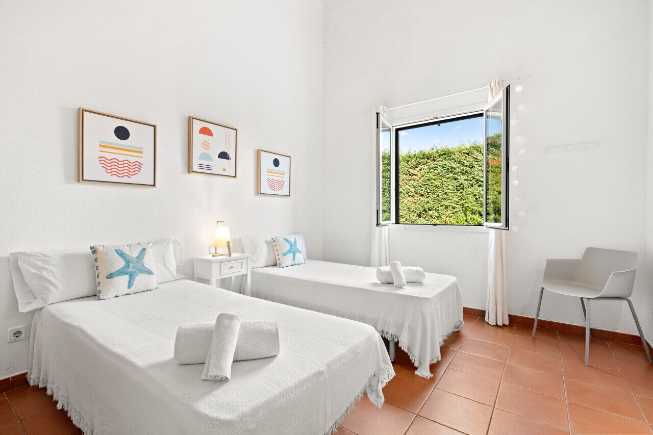 A hotel room with two beds, a window with a view of greenery, and a chair.