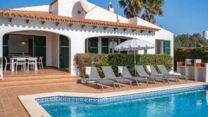 Una vila blanca amb teulada de rajola, piscina i zona de terrassa amb gandules i una taula.