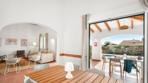 Un saló modern amb una taula de fusta, cadires blanques, un sofà i vista a una piscina i un jardí a través d'una porta oberta.