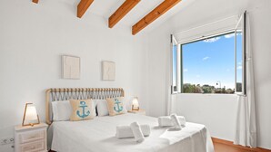 Un dormitori amb sostre de fusta, una finestra gran amb vistes, un llit amb roba de llit blanca i almoadins d'ancora, i una nitzeta amb una làmpada.