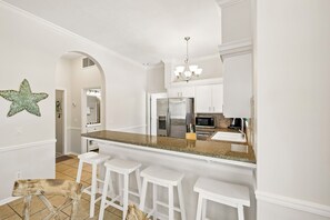 Eine Küche mit weißen Schränken, einem Edelstahl-Kühlschrank, einer Mikrowelle und einer Arbeitsplatte aus Granit. Drei weiße Barhocker und eine Meerstern-Dekoration sind an der Wand zu sehen.