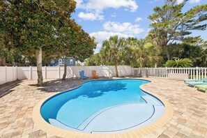 Ein Schwimmbecken mit Fliesen umgeben, umrahmt von einem weißen Zaun und Bäumen im Hintergrund.