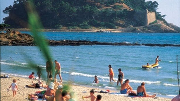 Ein Strand mit badenden und sonnenden Personen, ein Schloss auf einem Hügel im Hintergrund und ein klarer, blauer Himmel.