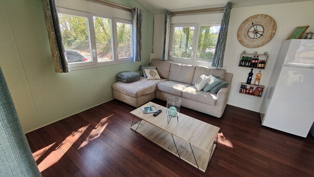Un salon avec un canapé beige, une table basse en bois et un réfrigérateur blanc. On y trouve des fenêtres avec des rideaux, une horloge au mur et une étagère avec des bouteilles.