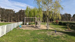 Une aire de jeux avec des balançoires et un toboggan, entourée d’une zone délimitée par une clôture et d’arbres.