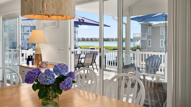 A dining area with a table, chairs, a vase with flowers, a lamp, and a view of the beach and water through a glass door. - Canal Front Views, Boat Dock, Kayaks, 10" Walk to the Beach, Free Activities inc. Golf at Bear Trap. (Fenwick Island)