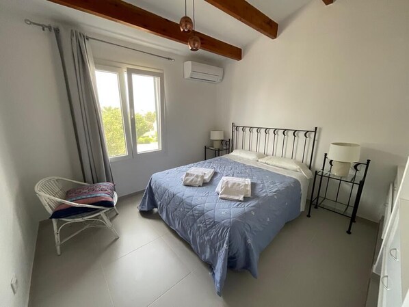 Un dormitorio con cama, dos mesitas de noche, una silla, una ventana y un equipo de aire acondicionado.
