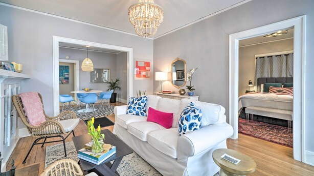 Un salon moderne avec un canapé blanc, des coussins aux motifs bleus et roses, un fauteuil en osier, une table basse et un lustre.