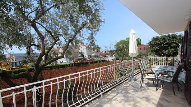 A balcony with a white metal railing, outdoor furniture, and a view of a landscaped area with trees and buildings in the background. - Apartment Paulinic (Rovinj)
