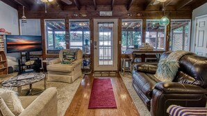 A cozy living room with wooden floors, leather sofas, a TV, and a large window overlooking a backyard.