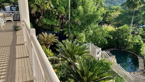 Um deck de madeira com guarda-corrimão branco, com vista para um jardim exuberante e uma piscina.