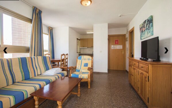 Una habitación con un sofá a rayas, una mesa de madera, una silla, un televisor sobre un mueble de madera, y una puerta con un letrero.