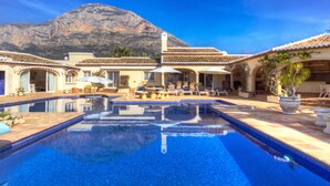Piscina com bordas azulejadas, cadeiras de espreguiçadeira e guarda-sóis, cercada por edifícios com telhados de azulejo e uma montanha ao fundo.
