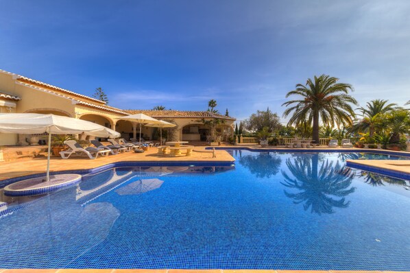A swimming pool with tiled edges, lounge chairs, umbrellas, and palm trees.