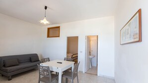 Un espace de vie compact comprenant un canapé, une table à manger, des chaises, un luminaire suspendu, un tableau encadré et une porte donnant accès à une salle de bain.