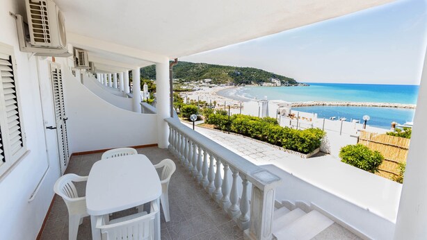 Un balcon avec une table et des chaises blanches, donnant sur une plage et la mer.