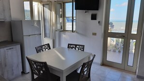 Un coin repas avec une table blanche et des chaises noires, un téléviseur à écran plat fixé au mur et une vue sur la mer grâce à de grandes fenêtres.