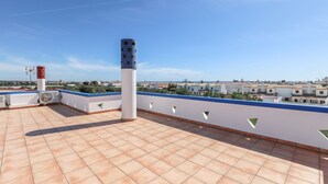 Una terraza en la azotea con suelo de baldosas, una pared blanca y azul con ventanas recortadas, y una vista despejada de edificios a lo lejos.