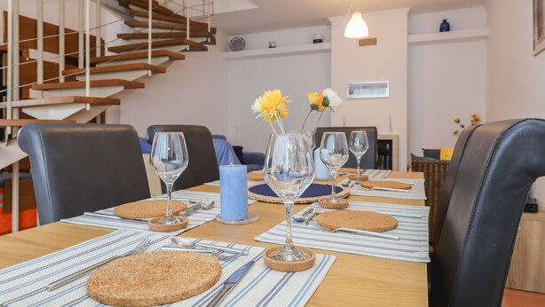 Un comedor con una mesa de madera dispuesta para cuatro personas, con individuales a rayas, posavasos redondos y vasos transparentes. En la mesa hay una vela azul y un florero con flores.