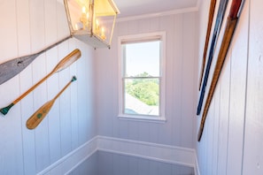 A narrow stairway with white paneled walls, a window with a view of greenery, and wooden paddles and skis hanging on the wall. - Beaulieu-Sur-Duck - Sanderling, Ocean View, Steps to Direct Boardwalk, Pets OK, Flex Stays (Duck)