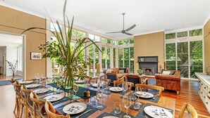 Une salle à manger avec une grande table dressée pour six personnes, entourée de chaises en bois. On aperçoit une cheminée au fond de la pièce, et celle-ci est dotée de grandes fenêtres.